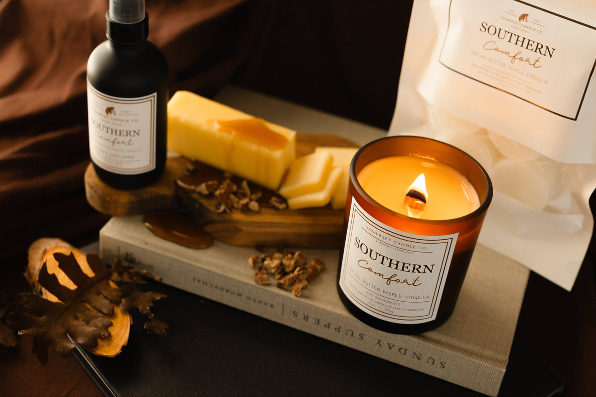 A styled product arrangement featuring a Southern Comfort candle in an amber glass jar with a lit flame, a dark bottle labeled Southern Comfort spray, a white bag of Southern Comfort wax melts, and a stick of butter on a book against a dark brown fabric backdrop.