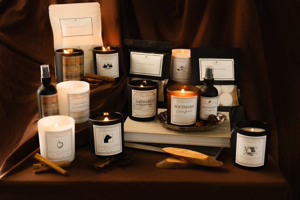 A styled product display of Somerset Candle Fall candles and related home fragrance items arranged on a draped dark brown fabric background. The set includes black glass jars with lit candles, white wax candles, dark bottles, and branded boxes and sachets, with cinnamon sticks and small decorative props on a table in front.