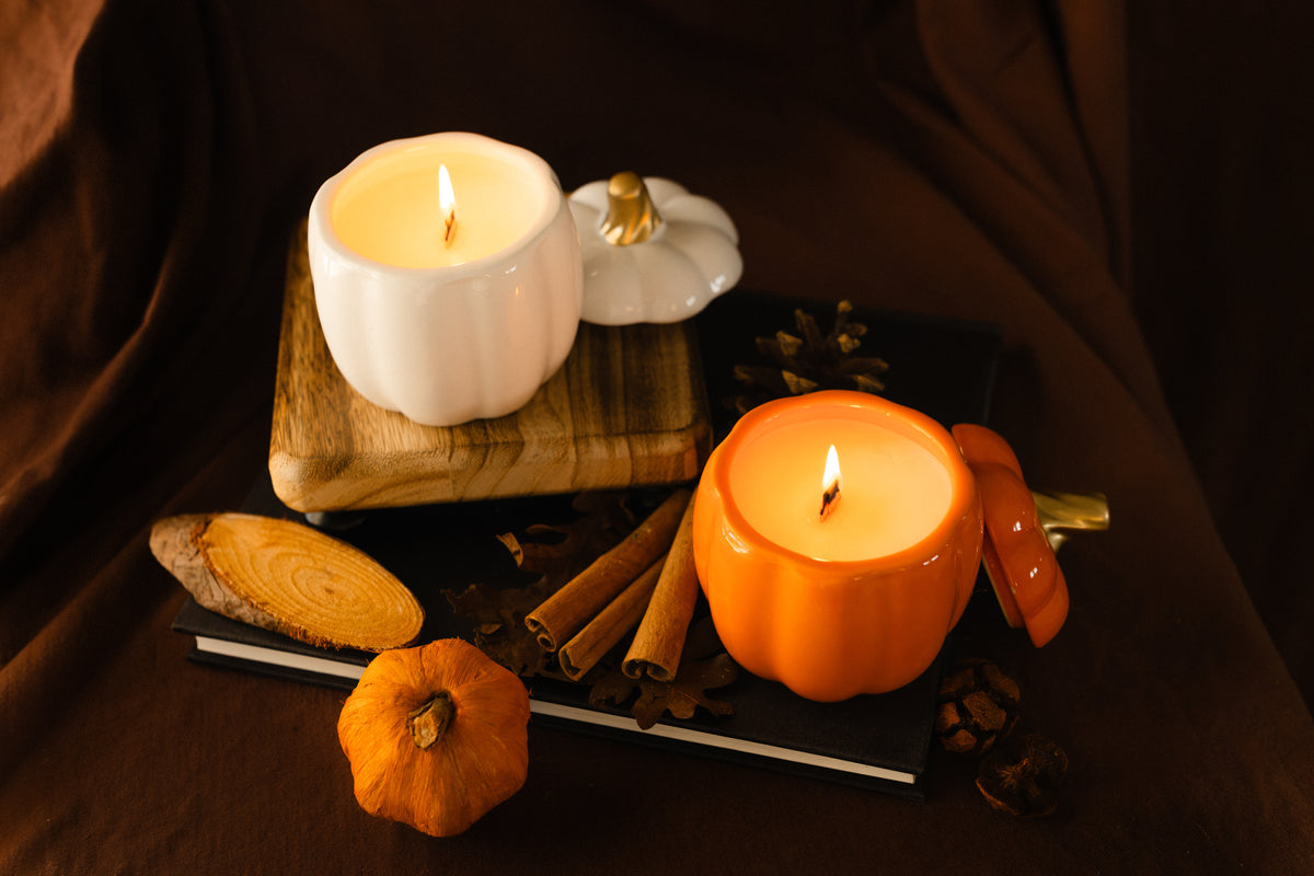 Two lit wax candles shaped like pumpkins, one white and one orange, styled on a dark cloth with cinnamon sticks, a small pumpkin, and wood slices.