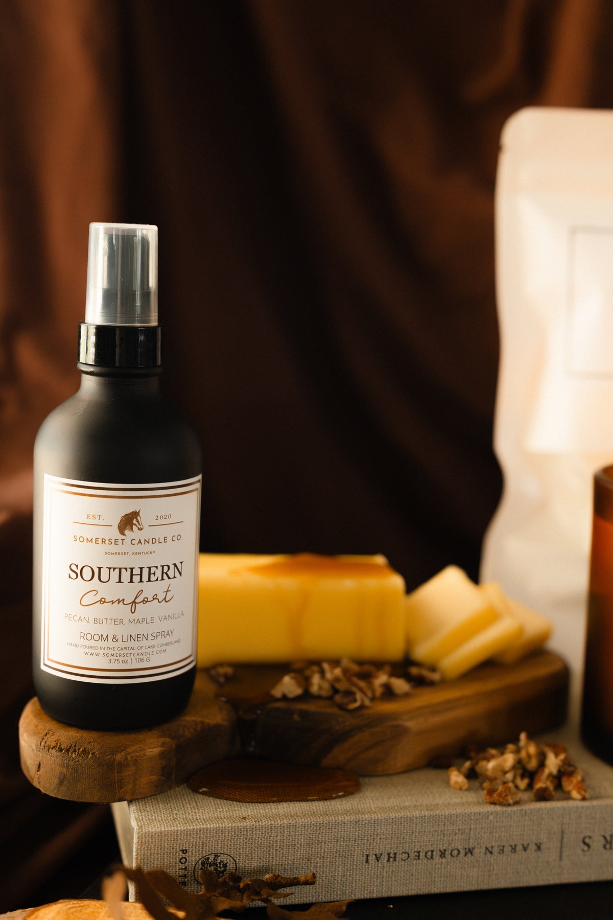 A black spray bottle labeled 'Southern Comfort' by Somersw Candle Co., styled on a wooden surface with cheese, dried herbs, and a book against a warm brown background.